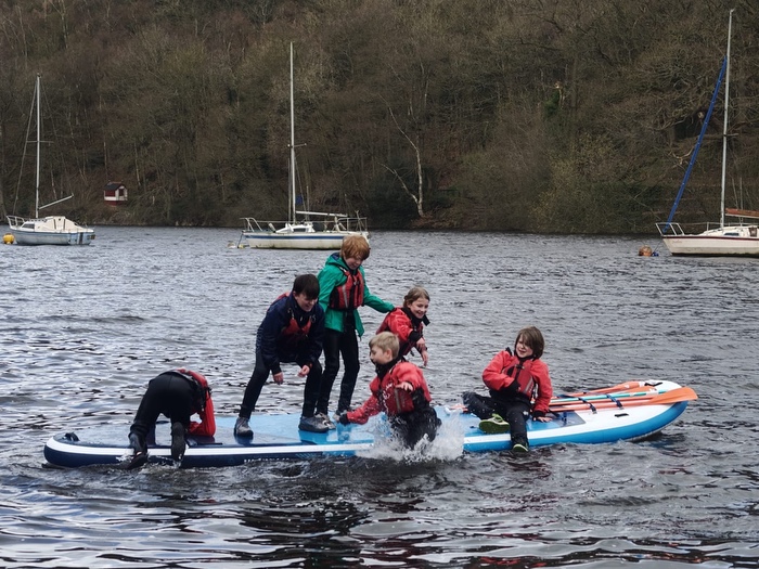 6 children in wetsuits messing about on a large stand up paddle board on a lake